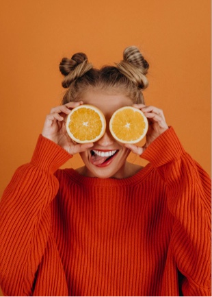 Sophie covering her eyes two orange slices, dressed in an orange sweater and standing in front of a orange background.