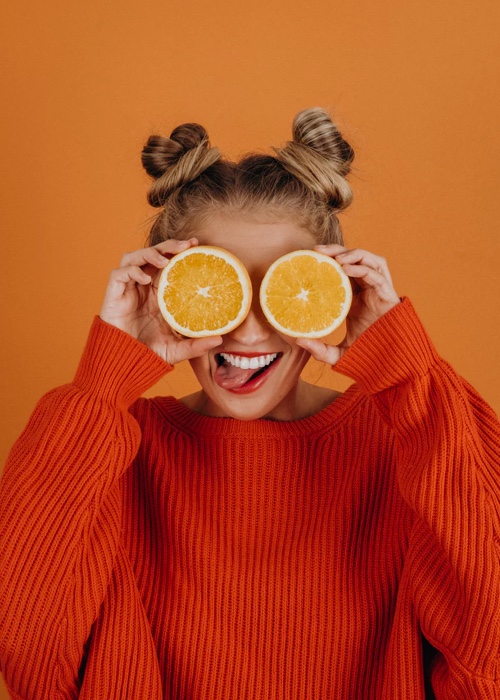 an image of Sophie, the blog creator, dressed in an orange sweater holding two slices of oranges in front of both her eyes standing in front of an orange background