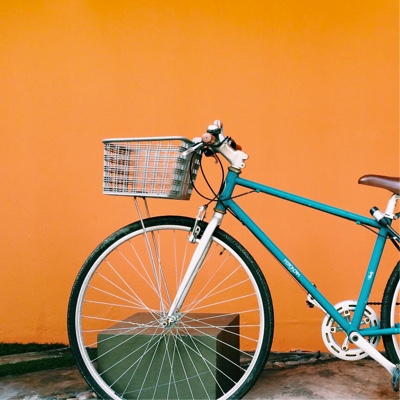 a photograph of a bicycle with a teal frame, white spokes and handle bars with a basket in the front to hold some items parked in front of a bright orange wall